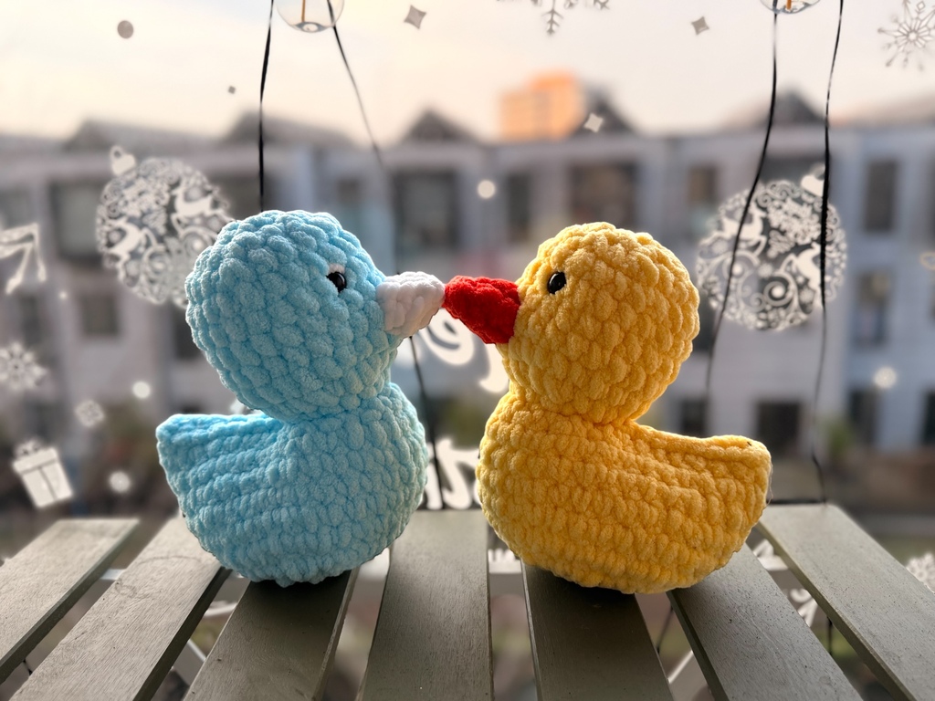 Two crochet ducks, one blue and one yellow with a red beak, on a railing with a blurred cityscape in the background.