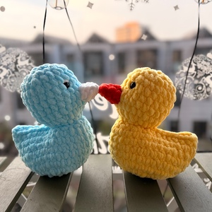 Two crochet ducks, one blue and one yellow with a red beak, on a railing with a blurred cityscape in the background.