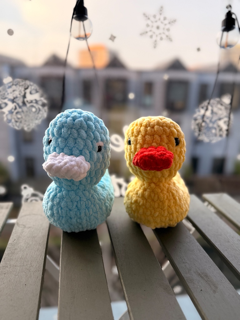 Two crochet ducks, one blue and one yellow with a red beak, on a railing with a blurred cityscape in the background.
